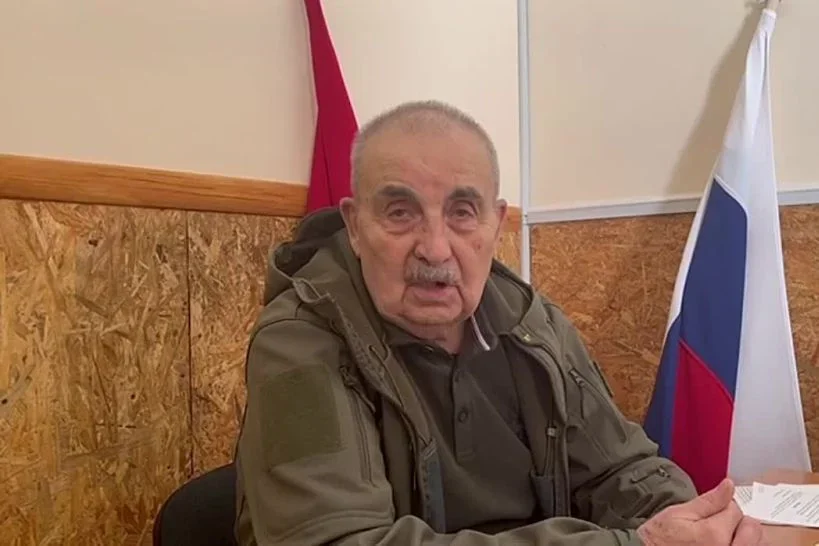 Літній чоловік у оливковій куртці сидить за столом на тлі російського прапора
