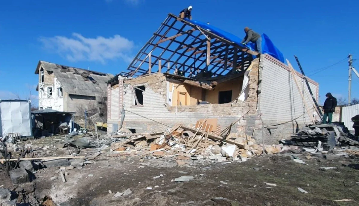 Пошкоджений цегляний будинок з відкритим каркасом даху, люди накривають його синім тентом серед уламків