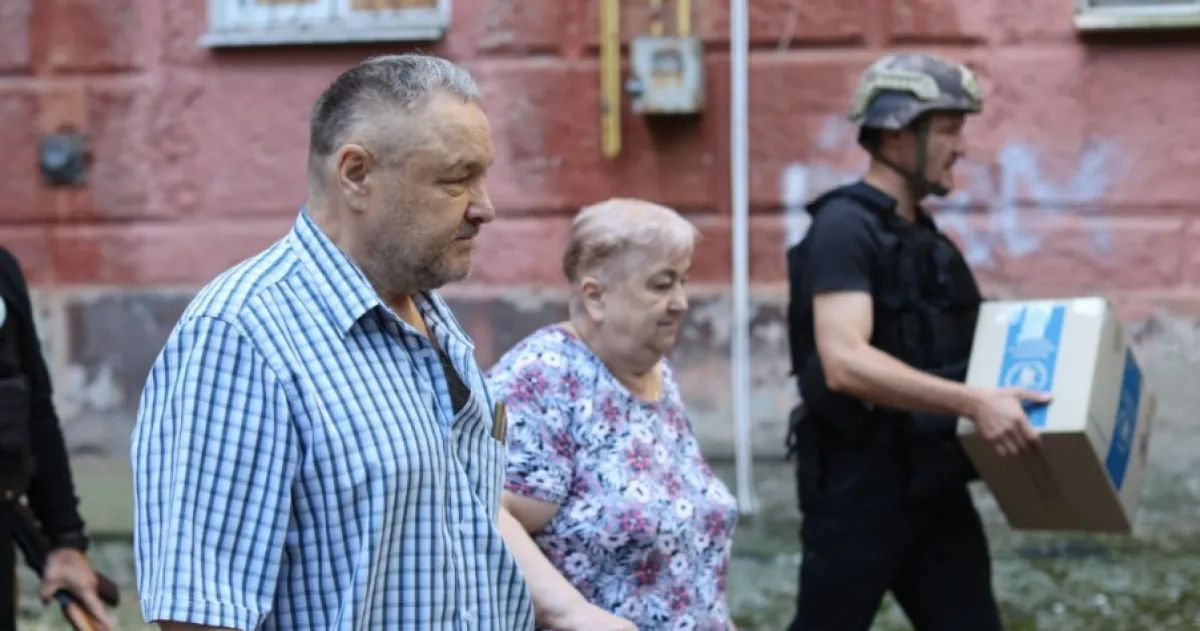 Літнє подружжя йде біля червоної стіни, військовий несе коробку